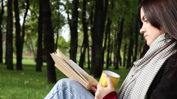 istock A woman sits under a tree and reads her favorite book while drinking coffee or tea from a yellow cup in a city park on green grass on a pleasant sunny day. Vacation, education and study concept 1288633145