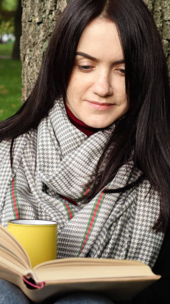 istock A woman sits under a tree and reads her favorite book while drinking coffee or tea from a yellow cup in a city park on green grass on a pleasant sunny day. Vacation, education and study concept 1288633173