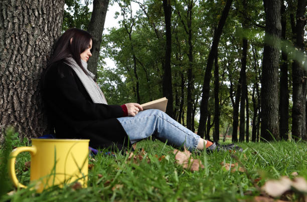istock A woman sits under a tree and reads her favorite book while drinking coffee or tea from a yellow cup in a city park on green grass on a pleasant sunny day. Vacation, education and study concept 1288633199