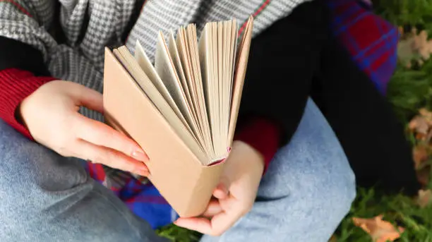 Photo of Girl reading a book in the autumn park. Female hands open the pages of a paper book outdoors on a warm sunny day. The student is preparing for the exam. Literary leisure in nature