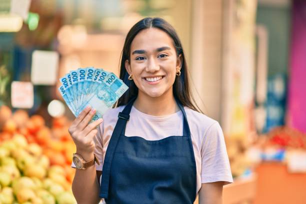 junge lateinische ladenbesitzer mädchen lächelnd glücklich halten brasilien echte banknoten im obstgeschäft. - real food fotos stock-fotos und bilder