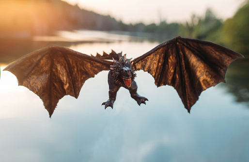 terrifying powerful dragon, Handmade mystical creature with strong wings, reptile with spikes and long tail, evil serpent with devilish look attacks enemy, magical animal flies over lake at sunset.