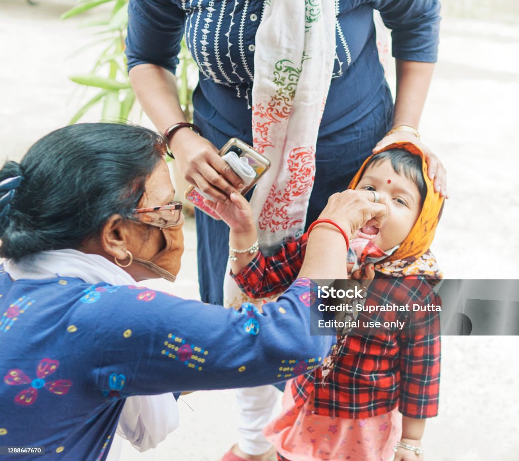 Pulse Polio vaccination programme in West Bengal West Bengal, 11-24-2020: A female ASHA (accredited social health activist) volunteer (the woman wearing protective face mask due to coronavirus pandemic) administering Pulse Polio vaccination to a cute little girl, accompanied by her mother. Polio Vaccine Stock Photo
