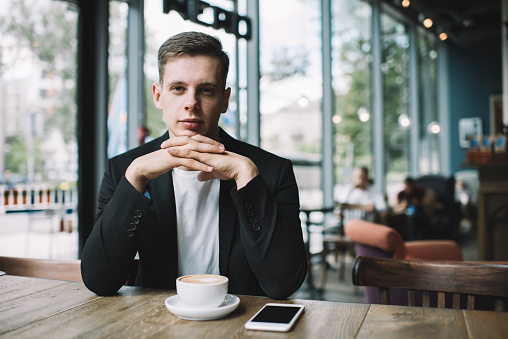 Confident young male with crossed fingers in black jacket and white t-shirt looking at camera with cup of cappuccino and smartphone on table in cafe
