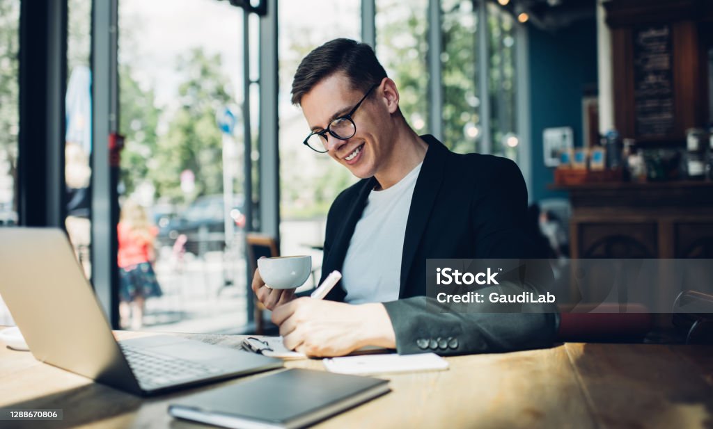 Happy businessman with cup of coffee writing notes Happy young male entrepreneur in eyeglasses and formal suit with cup of coffee sitting in cafe and writing notes while working on project Laptop Stock Photo