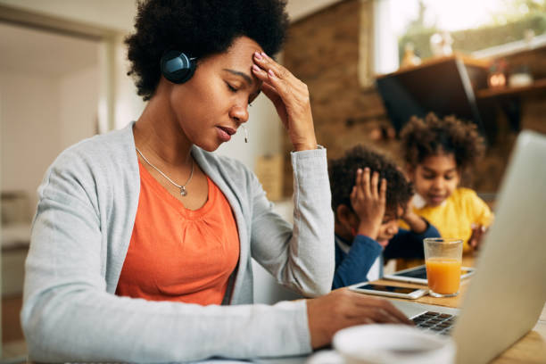 Black single mother feeling frustrated while working at home. African American mother feeling stressed out while working on laptop at home. Her kids are surfing the net on touchpad beside her. Tensed stock pictures, royalty-free photos & images