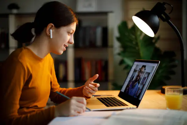 Photo of Smiling teenage girl with bluetooth headphones having video call on laptop computer at home