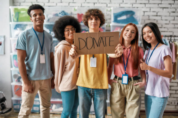 istock Group of diverse young volunteers smiling at camera while standing in charitable organization office, Guy holding card with Donate lettering 1288693978