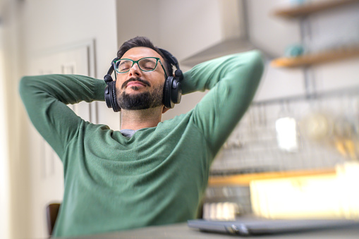 Man with hands behind head and eyes closed listening to music with headphones while sitting at dining table.