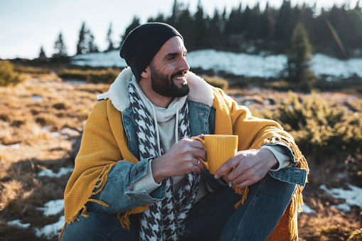 Handsome man drinks morning coffee, while sitting in forest on mountain, and enjoy fresh air on cold winter day