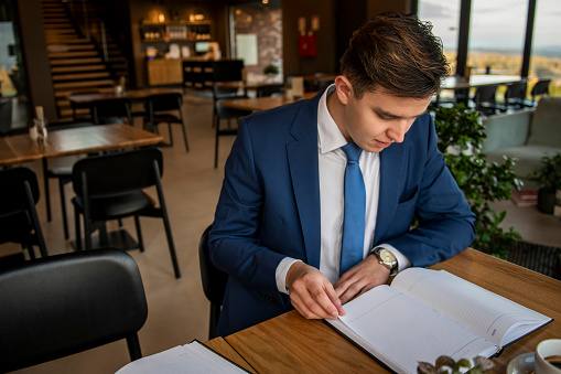 A young businessman is sitting in a cafe, he has coffee on the table and a planner - A businessman in an elegant suit with a tie is sitting thoughtfully in a cafe