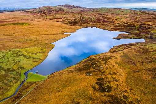 The view from a drone of a small Scottish loch that is used as a reservoir to supply water to a part of Dumfries and Galloway. The image was captured on an overcast autumn morning. The loch is in a remote rural area of south west Scotland. The reflection of the sky can be seen in the calm still water.