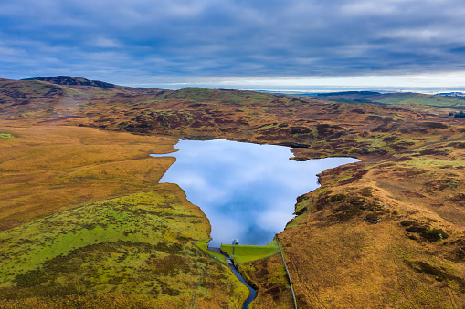 The view from a drone of a small Scottish loch that is used as a reservoir to supply water to a part of Dumfries and Galloway. The image was captured on an overcast autumn morning. The loch is in a remote rural area of south west Scotland. The reflection of the sky can be seen in the calm still water.