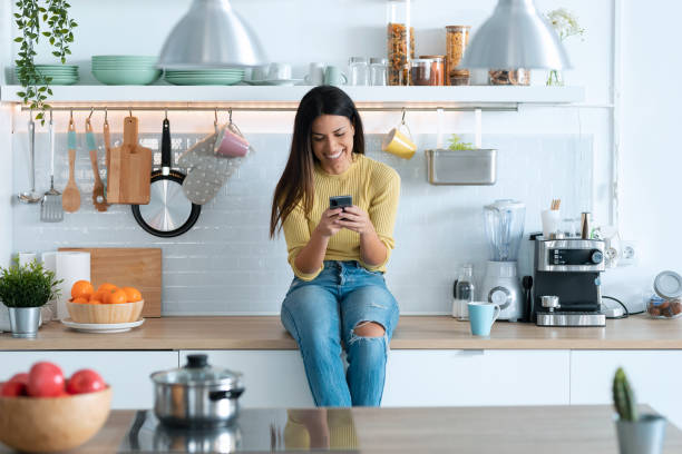 istock Attractive young woman using smart phone while relaxing sitting on the kitchen at home. 1288720251