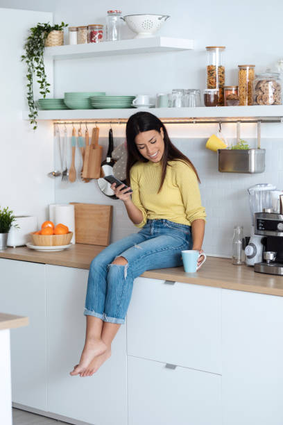 istock Attractive young woman using smart phone while relaxing drinking coffee sitting on the kitchen at home. 1288720343