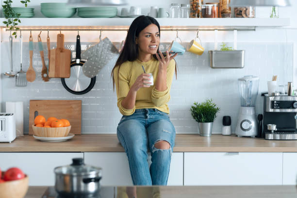 istock Attractive young woman eating yogurt while using her smart phone sitting on the kitchen at home. 1288720413