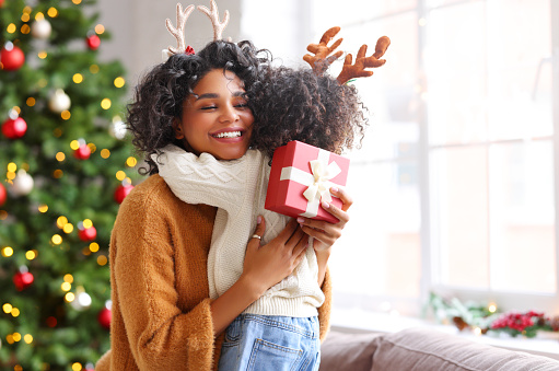 Cheerful ethnic woman with present smiling and embracing kid on sofa while celebrating Christmas at home