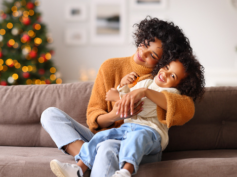 Cute ethnic boy in sweater playing and bonding with happy woman on comfortable couch on Christmas eve at home