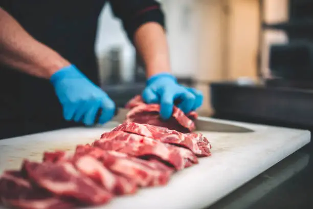 closeup view of a young caucasian man cutting raw pork neck with sharp knife. He makes steaks for grill in restaurant kitchen