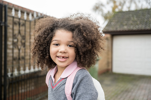 Head and shoulders view of 5 year old girl with curly brown hair wearing backpack and pausing to smile at camera on her way to school.
