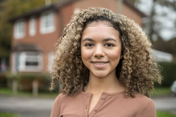 Portrait of cheerful mixed race teenage girl Head and shoulders view of 17 year old girl with curly brown hair wearing casual top and smiling at camera as she stands outdoors in residential district. 16 17 years stock pictures, royalty-free photos & images