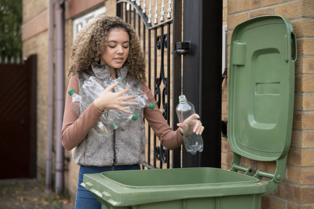 Responsible teenager putting plastic bottles in recycle bin Front view of 17 year old mixed race girl carrying armful of plastic bottles and placing them in recycling bin outside family home. hampshire england photos stock pictures, royalty-free photos & images