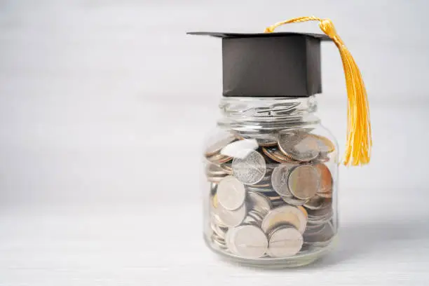 Photo of Graduation gap hat on coins money in jar for education fund; study learning concept.