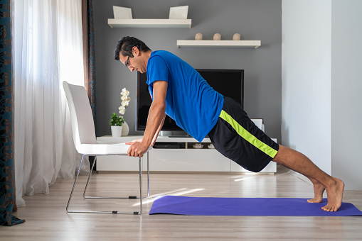 Young athletic man is doing push up exercise with chair at home in his spacious living room with modern interior. Gym at home