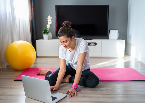 Young athletic beautiful woman is using laptop computer for watching online fitness live streaming classes on floor of living room at home. Gym at home