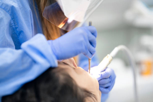Dental exam at dentist office Woman dentist with protective face mask, blue glove, and blue workwear. She is using dental equipment. Young patient opening her mouth and looking at dentist. dental drill stock pictures, royalty-free photos & images