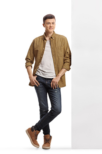 Full length portrait of a young man in shirt and jeans leaning on a wall isolated on white background