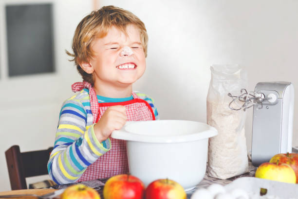 cute little happy blond preschool kid boy baking apple cake and muffins in domestic kitchen. funny lovely healthy child having fun with working with mixer, flour, eggs, fruits. little helper indoors - cake making mixing eggs imagens e fotografias de stock