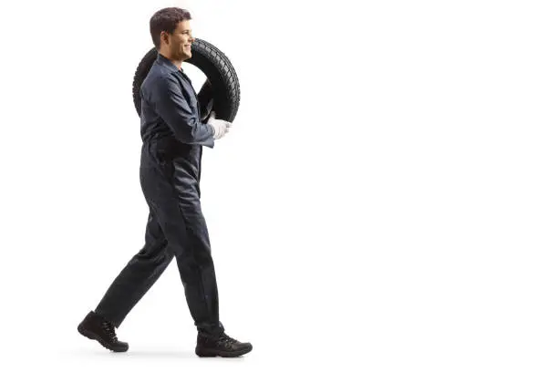 Full length profile shot of an auto mechanic walking and carrying a tire on his shoulder isolated on white background