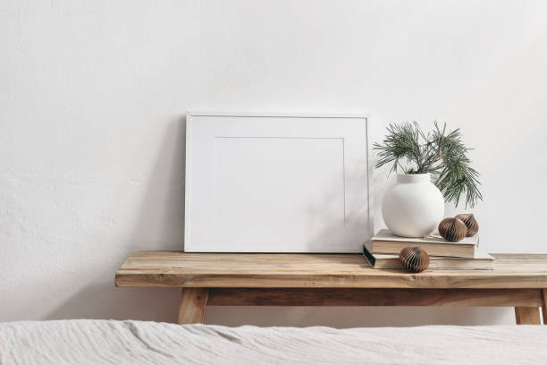 istock Winter still life. Horizontal white frame mockup on vintage wooden bench, table. Modern white ceramic vase with pine tree branches, Christmas paper ornaments and books. White wall background. 1288795048