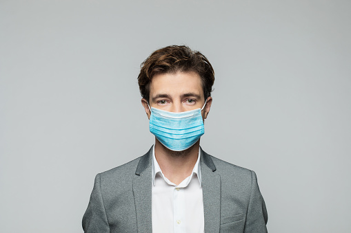 Portrait of confident young businessman wearing N95 face mask looking at camera. Studio shot, grey background.