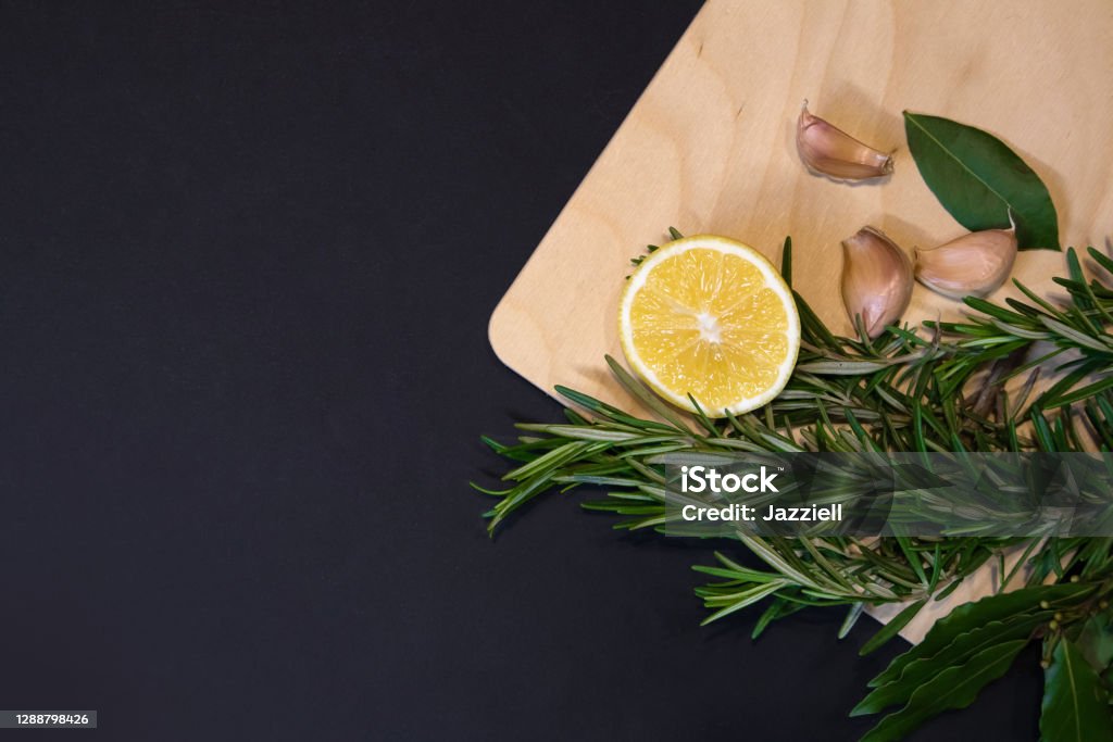Cutting board with bay leaves, rosemary sprigs, lemon and cloves of garlic Cutting board of light wood framed with bay leaves, rosemary sprigs, lemon and cloves of garlic, designer template with dark gray background Bay Leaf Stock Photo