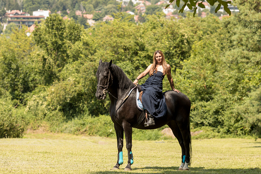 Girl with a long copper hair and long flowy shoulderless dress ride a black horse with trees in the background.