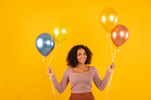Smiling young woman holding balloons while standing against yellow background