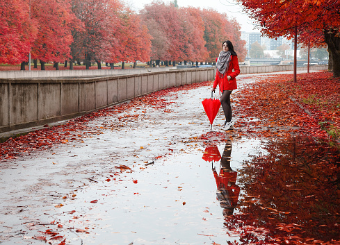 young girl in a red coat with an umbrella stands on the alley of the park after the rain on an autumn day