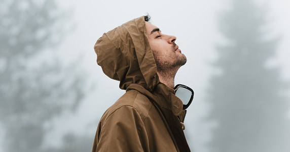 Side view of a handsome male walking in misty nature, listening to the music on his headphones with closed eyes outside. Traveler man wearing a stylish coat in the mountain enjoying the fresh air.