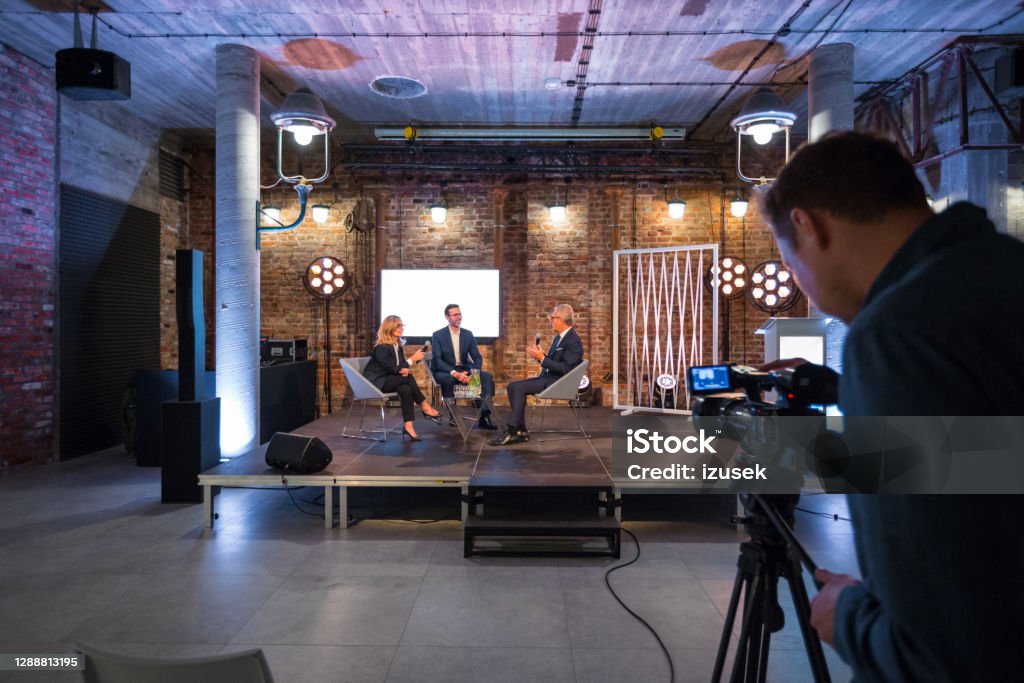 Business people during video conference Businesswoman and businessman giving presentation during online seminar, sitting on armchairs on the stage, holding microphones. Video camera on the foreground. Conference - Event Stock Photo