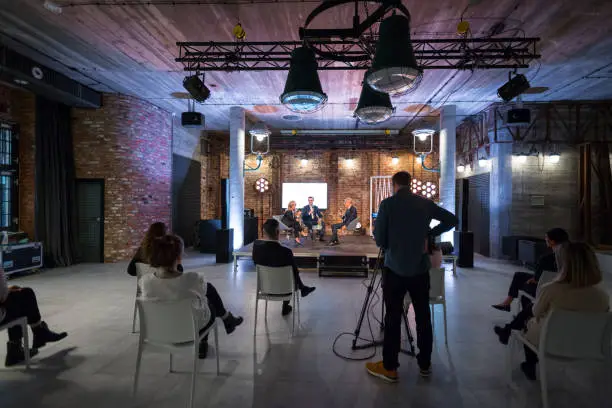 Business people giving presentation during online seminar, sitting on armchairs on the stage, holding microphones. Audience sitting on chairs, keeping distance.