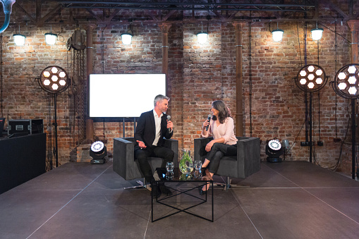 Businesswoman and businessman giving presentation during seminar, sitting on armchairs on the stage, holding microphones. Wide angle view.