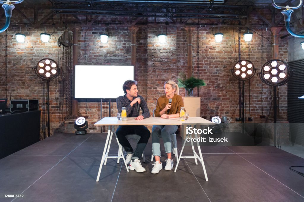 Young adult people during discussion in desing studio Young adult woman and man giving presentation during seminar, sitting at the table on the stage, holding microphones and discussing. Wide angle view. Interview - Event Stock Photo