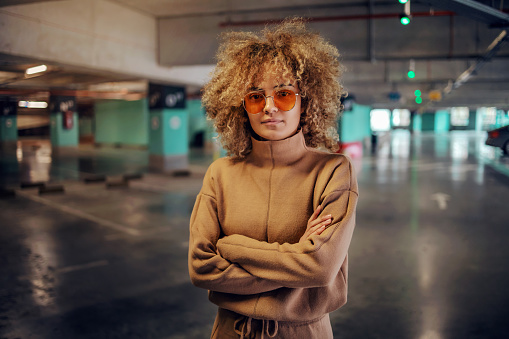Young fashionable woman with curly hair standing in underground garage with arms crossed and looking at camera.