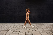 Young fashionable woman with curly hair walking on the street and listening to the music.