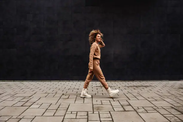 Young fashionable woman with curly hair walking on the street and listening to the music.