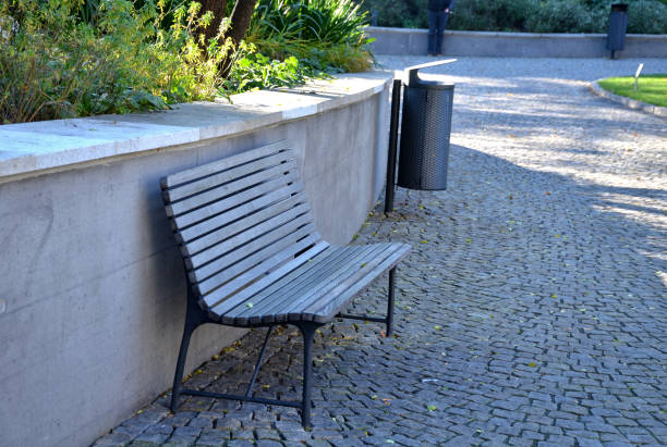 un banc avec un siège lambrissé gris par le mur de soutènement sur un trottoir de cube de granit dans un parc de ville et une poubelle en métal - footpath autumn stone old photos et images de collection