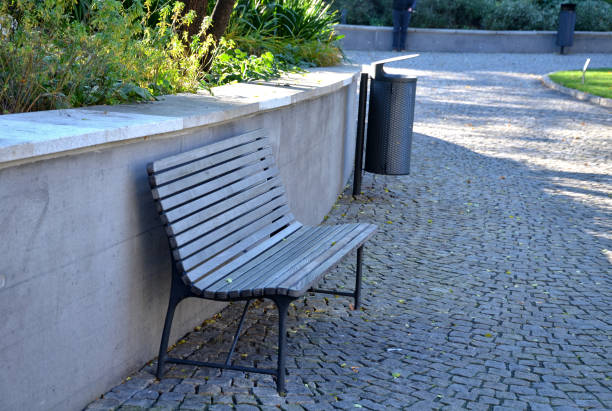 скамейка с серым панельным сиденьем у подпорной стены на гранитном тротуаре куба в городском парке и металлический мусорный бак - brick yellow road footpath стоковые фото и изображения