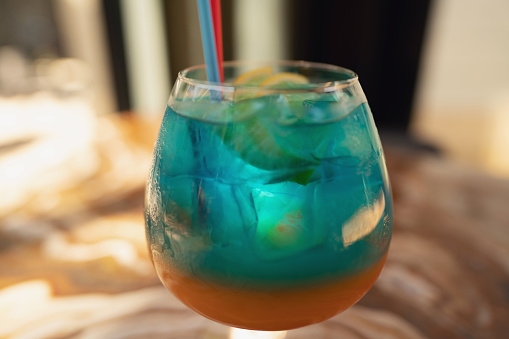 Close up photo of blue and orange drink in cocktail glass with lemon slices standing on marble bar counter. Focus on foreground.